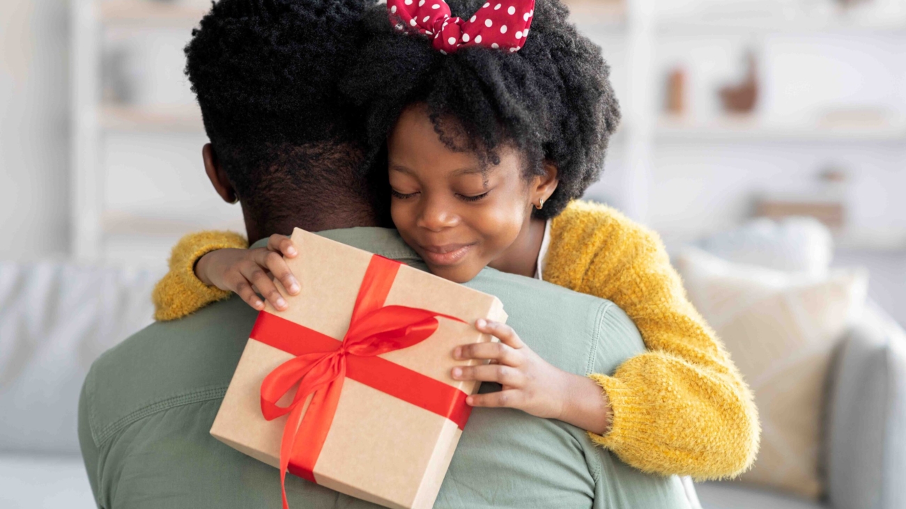 Smiling little black girl holding gift box and embracing dad, loving daughter greeting daddy with Father's day or birthday, happy african american family bonding together at home, closeup shot